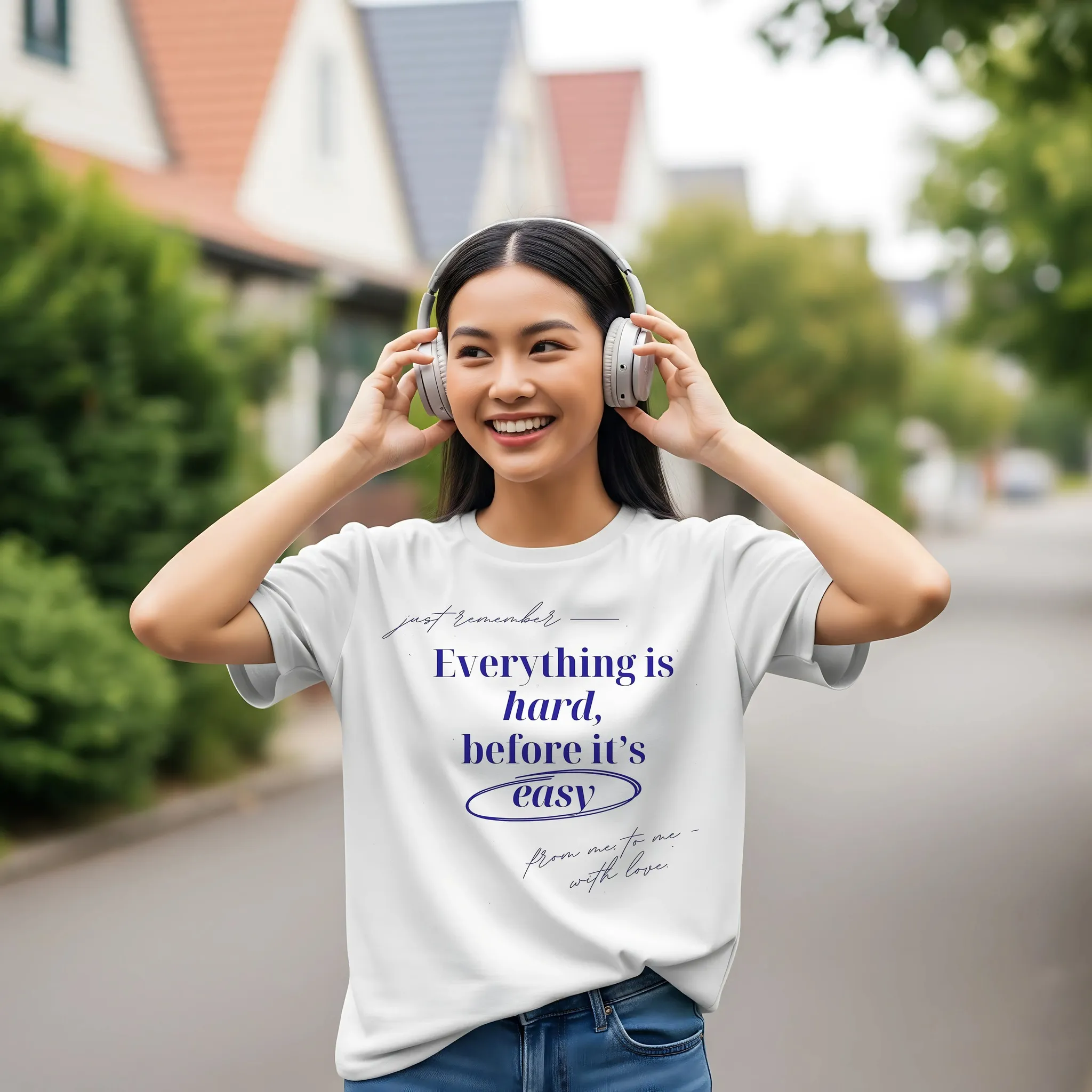 Smiling young woman wearing Nielseun white t-shirt with motivational quote "Everything is hard before it’s easy" while listening to headphones outdoors
